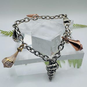 May include: A silver chain link bracelet featuring multiple seashell charms in various metallic finishes, including polished silver, copper, and gold tones. The charms are suspended from the circular links, which are secured with a lobster clasp.