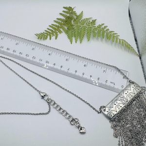 May include: A silver-toned necklace featuring a rectangular pendant with etched scrollwork and a long fringe of fine chains. The necklace includes a lobster clasp and an extender chain with a small heart charm, measured by a ruler in inches.