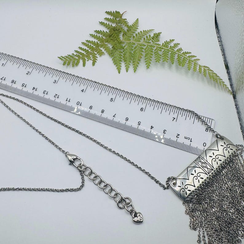 May include: A silver-toned necklace featuring a rectangular pendant with etched scrollwork and a long fringe of fine chains. The necklace includes a lobster clasp and an extender chain with a small heart charm, measured by a ruler in inches.