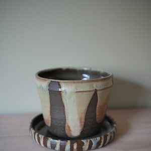 May include: A handmade ceramic planter with a matching saucer. The planter is a muted beige with dark brown drips and a dark brown rim. The saucer has a striped pattern in brown, beige, and blue. The set is suitable for small plants.