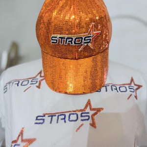 May include: An orange sequined baseball cap with the word "STROS" in white and blue, and a star logo. The cap is paired with a white shirt featuring the same logo and star design.