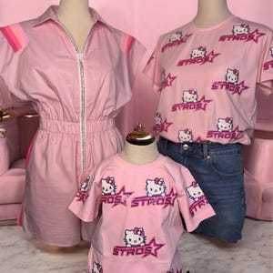 May include: Pink clothing set featuring a romper with a zipper and a t-shirt with a Hello Kitty design and the word "STROS". The t-shirt is paired with denim shorts. The set is displayed on mannequins.