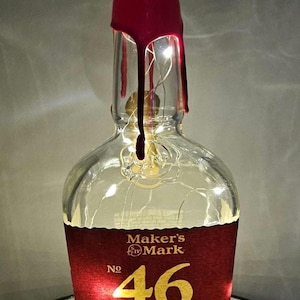 May include: A Maker's Mark bourbon whiskey bottle with a red wax seal. The label reads "No 46" and "Kentucky Straight Bourbon Whisky Barrel". The bottle is illuminated with fairy lights.
