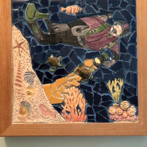 May include: A framed mosaic artwork depicting a scuba diver in a purple wetsuit with a green tank, surrounded by coral, seashells, and fish on a blue background.