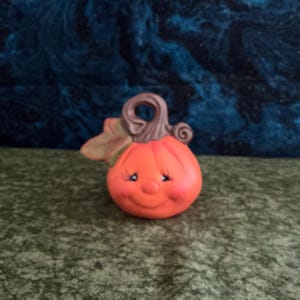 May include: A ceramic pumpkin figurine with a smiling face, rosy cheeks, and closed eyes. The pumpkin is orange with a brown stem and a green leaf. This decorative item is perfect for fall or Halloween.