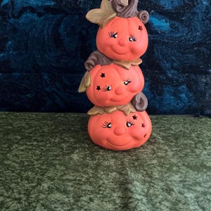May include: A stacked pumpkin figurine with a smiling face, star cutouts, and a brown stem. The orange ceramic decoration has a whimsical design, perfect for fall or Halloween.