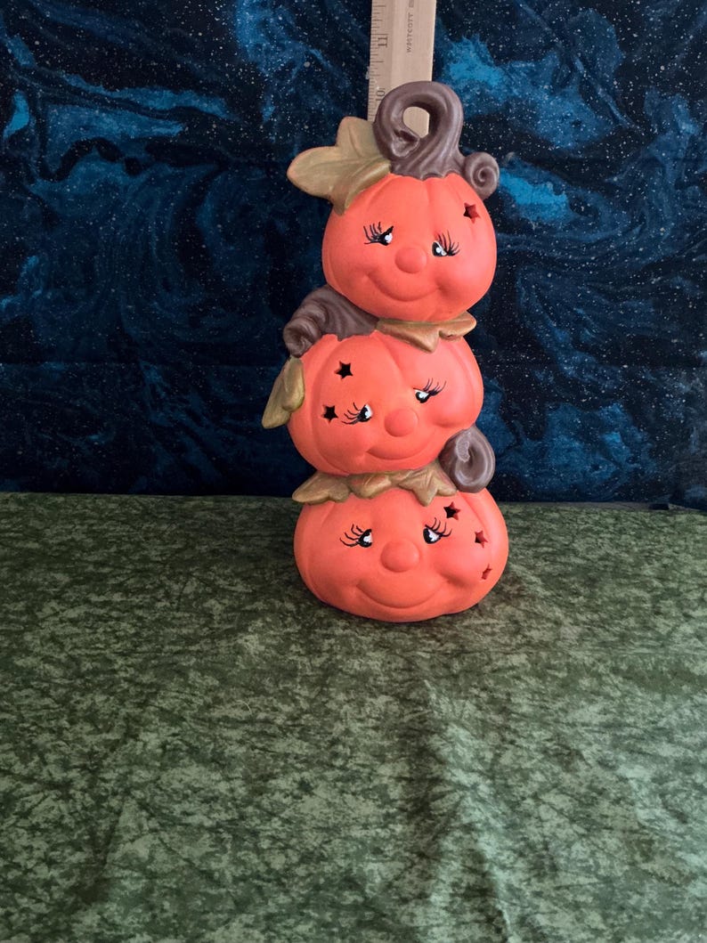 May include: A stacked pumpkin figurine with a smiling face, star cutouts, and a brown stem. The orange ceramic decoration has a whimsical design, perfect for fall or Halloween.