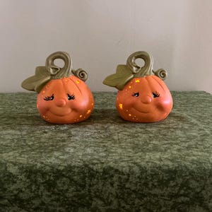 May include: Two orange ceramic pumpkin figurines with smiling faces, eyelashes, and star accents. Each pumpkin has a green stem and leaf. The pumpkins are displayed on a green textured surface.