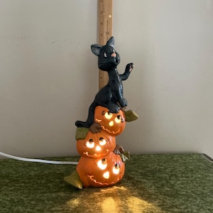 May include: A black cat figurine perched atop a stack of three illuminated orange jack-o'-lanterns. The ceramic sculpture features carved facial expressions and green leaf accents. A wooden ruler behind the object shows a height of 14 inches.
