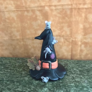 May include: A whimsical Halloween decoration featuring a black witch's hat with an orange band and buckle. The hat is adorned with small white mice, a purple berry, and autumn leaves. The base is black, and the overall design is charming.