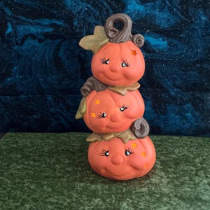 May include: A stacked pumpkin figurine with three smiling pumpkin faces, each with eyelashes and star accents. The pumpkins are orange with brown stems and leaves, set against a dark blue and green background. A whimsical Halloween decoration.