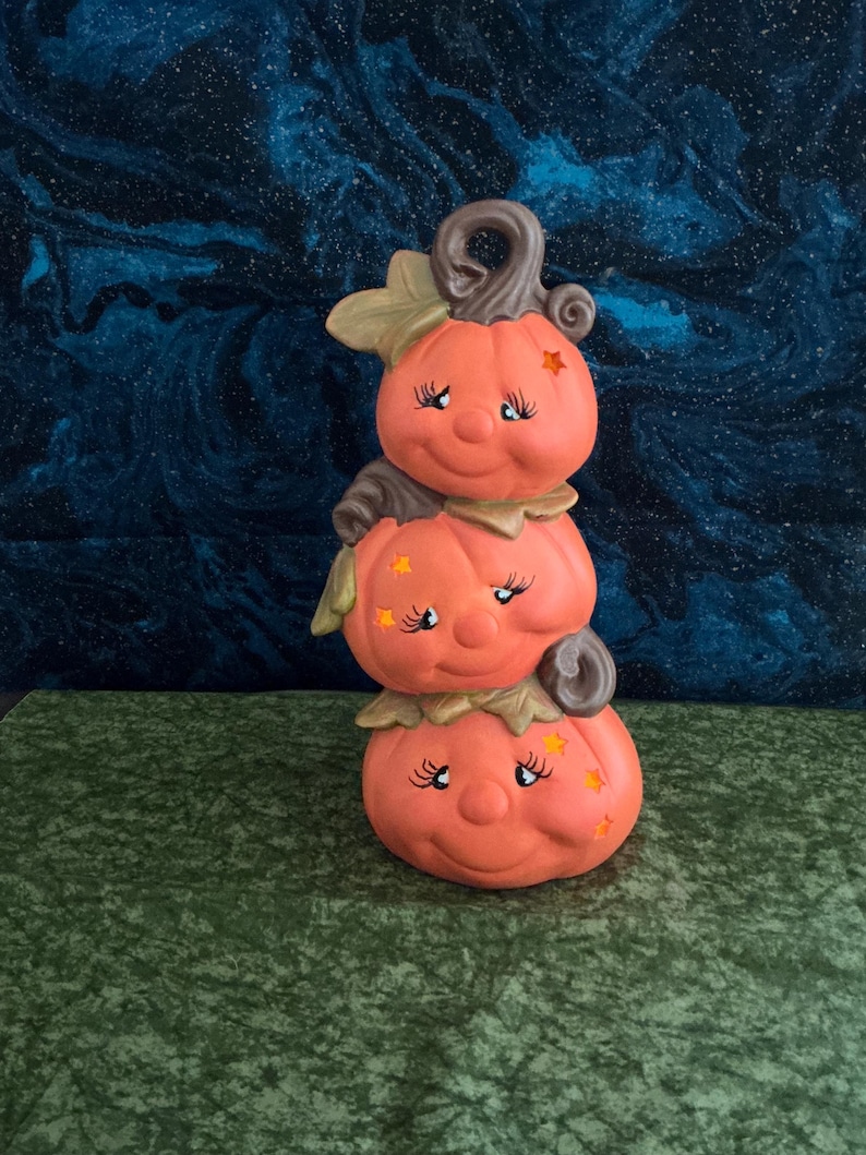 May include: A stacked pumpkin figurine with three smiling pumpkin faces, each with eyelashes and star accents. The pumpkins are orange with brown stems and leaves, set against a dark blue and green background. A whimsical Halloween decoration.