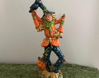 Puede incluir: Una figura decorativa de espantapájaros con sombrero azul, chaqueta naranja y pantalones azules con detalles de costuras amarillas. La figura sostiene un cuervo negro en una mano levantada y se apoya sobre una base con una calabaza naranja.