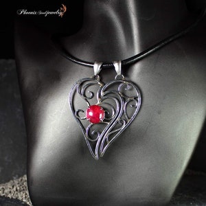 Può includere: Una collana con un ciondolo a forma di cuore in filigrana d'argento con una pietra preziosa rossa al centro. La collana è su un cordoncino in pelle nera.
