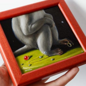 Op de afbeelding: Een klein ingelijst schilderij van een aap die op een groene ondergrond zit. De aap draagt een gouden ring aan zijn vinger en er liggen verschillende kleine rode en witte kralen rondom. Het schilderij is rechtsonder getekend met "vanitas".