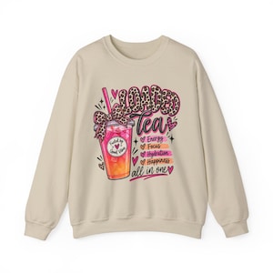 May include: A beige crewneck sweatshirt featuring a graphic of a pink and orange iced drink with a leopard print bow and straw. The text reads "LOADED Tea" with "Energy", "Focus", "Hydration", "Happiness", and "all in one" listed below.