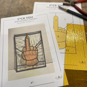 Op de afbeelding: Afbeelding van een glas-in-loodpatroon met de tekst "F*CK YOU". Het patroon toont een hand met de middelvinger omhoog. Ook zichtbaar zijn gereedschappen en glasstukken voor glas-in-loodkunst.