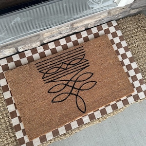 Peut inclure: Un paillasson en fibre de coco brun avec un motif abstrait noir. Le paillasson est pos&eacute; sur un tapis &agrave; carreaux.