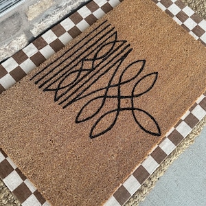 Peut inclure: Un paillasson en fibre de coco marron avec un motif g&eacute;om&eacute;trique noir. Le paillasson est pos&eacute; sur un tapis &agrave; carreaux brun et blanc.