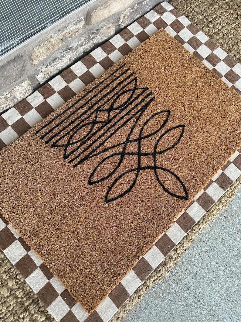 Peut inclure: Un paillasson en fibre de coco marron avec un motif g&eacute;om&eacute;trique noir. Le paillasson est pos&eacute; sur un tapis &agrave; carreaux brun et blanc.
