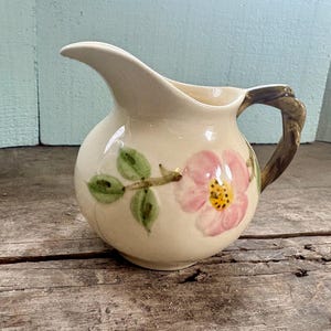 Puede incluir: Jarra de cerámica color crema con diseño floral. La jarra presenta una flor rosa con centro amarillo y hojas verdes. El asa es marrón y texturizada.