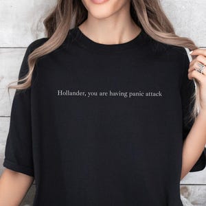 May include: Black t-shirt with the text "Hollander, you are having panic attack" in white. The shirt is worn by a person. The background is a light-coloured, wooden wall.