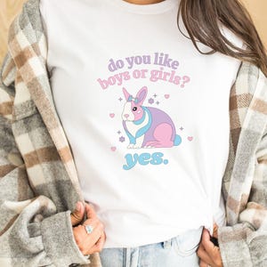 Könnte beinhalten: Weißes Langarm-T-Shirt mit einem rosa und blauen Comic-Kaninchen, das eine Schleife trägt. Der Text "do you like boys or girls? yes." ist auf das Shirt gedruckt.