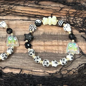 May include: Two beaded bracelets with a Halloween theme. One bracelet features black and white floral beads, black beads, and a clear ghost charm. The other bracelet has black and white checkered beads, a black star, and a yin-yang charm.