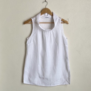 May include: A sleeveless white blouse with a high, rounded collar. The blouse is hanging on a wooden hanger against a neutral background. The fabric appears to be lightweight and slightly textured.