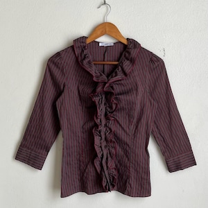 May include: A brown striped blouse with red accents, featuring a ruffled collar and front. The three-quarter sleeves and button-down front add to the blouse's design. The blouse is hanging on a wooden hanger.