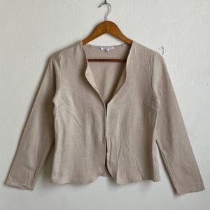 May include: A beige, long-sleeved cardigan with a round neckline and button closure. The cardigan is made of a textured fabric and hangs on a wooden hanger against a white wall.