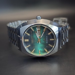 May include: A vintage wristwatch with a silver case and strap. The watch face has a green gradient with gold hands and markers. The day and date are displayed at the top. A classic timepiece, likely from the 1970s.