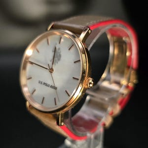 May include: A gold-toned wristwatch with a light-colored face and a brown leather band. The watch face displays the text "U.S. POLO ASSN." and has gold-colored hands and markers. The band has a red accent.