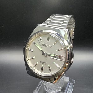Può includere: Orologio Orient color argento con quadrante grigio chiaro e cinturino in acciaio inossidabile. L'orologio presenta un display giorno e data ed è esposto su un supporto trasparente su sfondo scuro. Sul quadrante è stampata la parola "Orient".