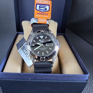 Op de afbeelding: Een zwarte Seiko 5 Sports automatische horloge met een zilveren bezel en een zwarte wijzerplaat. Het horloge heeft een datumvenster en een zwarte rubberen band. Het horloge zit in een doos met een Seiko 5 Sports label.