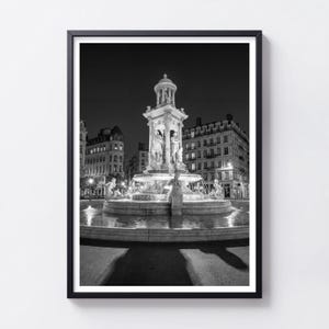 May include: A black and white framed wall art print featuring a night scene of a multi-tiered stone fountain with classical statues. The fountain is illuminated against a dark sky, surrounded by multi-story urban buildings with lit windows.
