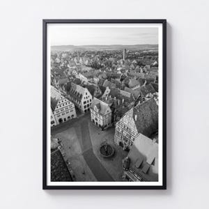 Può includere: Una stampa artistica da parete incorniciata in bianco e nero che mostra una vista aerea di una storica piazza cittadina europea. La scena presenta diversi edifici con tetti a capanna, architettura a graticcio e una fontana centrale.