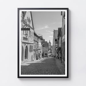 Può includere: Una stampa artistica da parete incorniciata in bianco e nero che raffigura una stretta strada acciottolata in una storica città europea. La scena presenta edifici a più piani con architettura tradizionale, un'insegna in metallo e una torre.