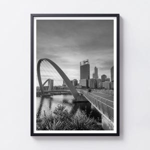 May include: A black and white framed wall art print featuring the Elizabeth Quay Bridge in Perth, Australia. The image captures the curved steel structure of the pedestrian bridge spanning over water with the city skyline visible in the background.