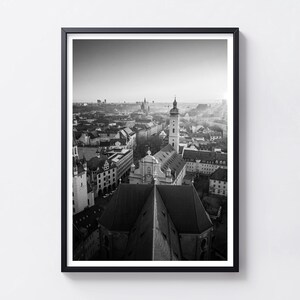 Pode incluir: Uma impressão emoldurada a preto e branco com uma vista elevada da cidade. A composição destaca o telhado de uma grande igreja em primeiro plano, guiando o olhar para uma torre de relógio alta e uma densa paisagem urbana ao fundo.