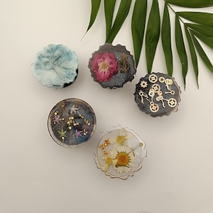 Könnte beinhalten: Vier runde Handygriffe aus Harz mit verschiedenen Designs. Einer hat ein blaues und weißes Marmormuster, einer eine rosa Blume, einer goldene Zahnräder und einer eine weiße Blume mit goldenen Akzenten.