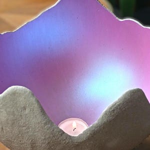Könnte beinhalten: Ein schalenförmiger Kerzenhalter mit einer rauen, strukturierten grauen Außenseite und einer glatten, irisierenden Innenseite, die zwischen leuchtendem Violett und sanftem Blau changiert. Ein kleines, brennendes weißes Teelicht steht am Boden.