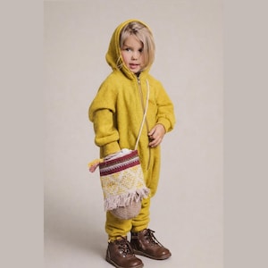 May include: A child wearing a mustard yellow hooded jumpsuit with a front zipper and brown leather boots. The child is carrying a small, patterned shoulder bag with a tassel. The bag features red, white, and yellow accents.