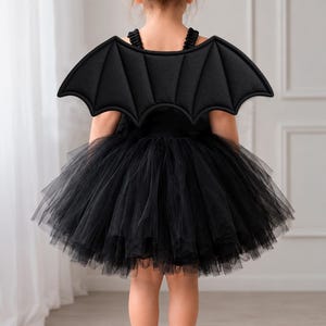 May include: A black dress featuring a layered tulle skirt and attached bat wings on the back. The bodice is solid black with shoulder straps, and the skirt consists of multiple tiers of sheer black tulle fabric. The bat wings are black, padded, and shaped like stylized bat wings.