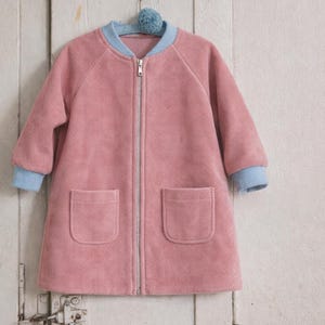 May include: A pink corduroy jacket with a light blue collar and cuffs, featuring a full-length zipper and two front pockets. The jacket is hanging on a blue pom-pom hanger against a white wooden door.