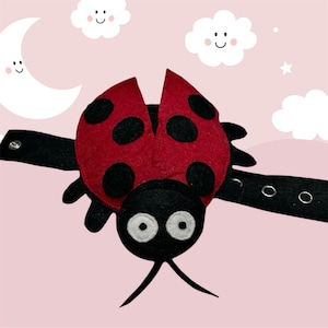 May include: A red and black ladybug-shaped accessory with black spots and a black strap. The ladybug has large white eyes and antennae. The strap has metal eyelets for adjustment. The background is pink with cartoon clouds, a moon, and stars.