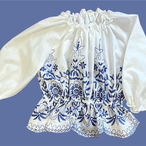 May include: White peasant top with blue floral embroidery. The top has a ruffled neckline and long sleeves.