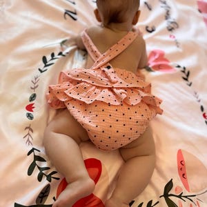 May include: A peach-colored baby romper with small black polka dots, featuring ruffled layers on the back and crisscross shoulder straps. The garment is made of a textured fabric and is worn by an infant lying on a patterned blanket.