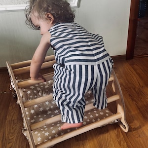 May include: A wooden climbing triangle with a beige fabric cover featuring a white star pattern. The structure consists of two side panels with arched cutouts connected by horizontal wooden rungs, designed for indoor physical activity.