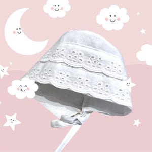 Puede incluir: Gorro de bebé blanco con delicado ribete de encaje floral y cierre de lazo. El gorro está hecho de una tela suave y presenta una corona redondeada y un ala ancha. El fondo es de color rosa claro con ilustraciones de luna, nubes y estrellas.